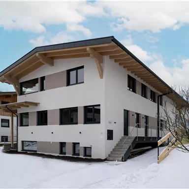 Ein modernes Wohnhaus in schneebedeckter Landschaft. Die Fassade ist weiß mit großen Fenstern und einem Holzüberstand.