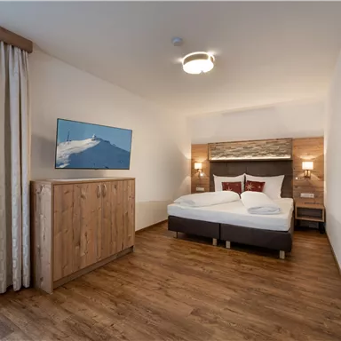 Ein modernes Zimmer mit einem Doppelbett und Holzfußboden. Ein Fernseher ist an der Wand angebracht, und es gibt Vorhänge, die ein wärmeres Ambiente schaffen.