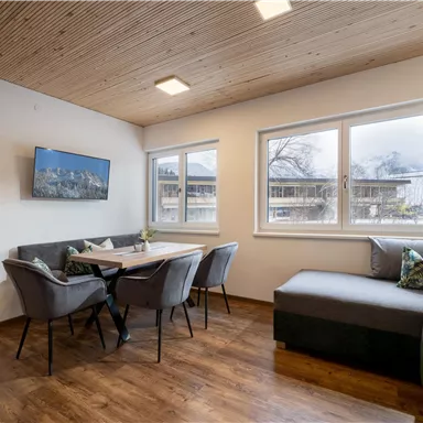 Ein modernes Wohnzimmer mit einem gemütlichen Sofa und einem Essbereich. Große Fenster sorgen für viel Tageslicht.