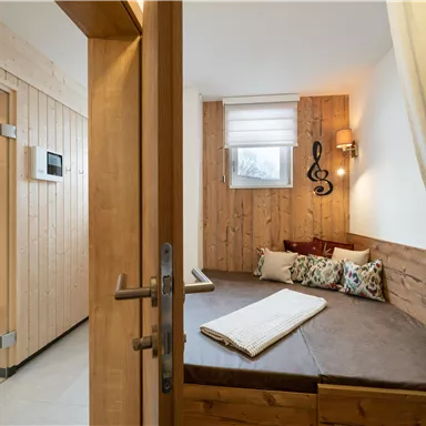 Ein gemütlicher Wellnessbereich mit einem Holzsofa und einer Sauna im Hintergrund. Die Wände sind aus Holz, was eine entspannende Atmosphäre schafft.