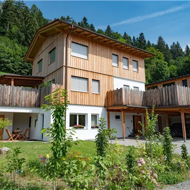 Ein modernes Holzhaus mit einem großen Balkon und einem gepflegten Garten. Im Hintergrund sind grüne Hügel und Bäume sichtbar.