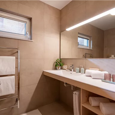 Ein modernes Badezimmer mit hellem design und Holzoberflächen. Es sind Handtücher und Pflegeprodukte auf der Ablage sichtbar.