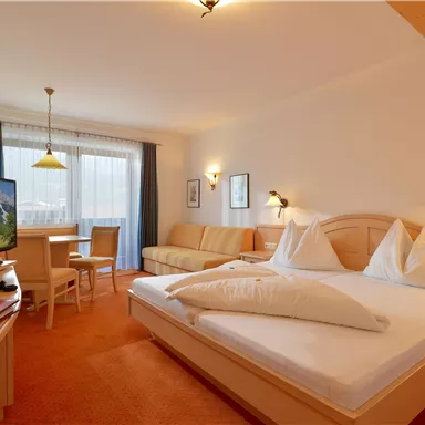 Ein gemütliches Hotelzimmer mit einem Doppelbett und einem Fernseher. Es gibt einen Tisch und Stühle sowie eine große Fensterfront mit natürlichem Licht.