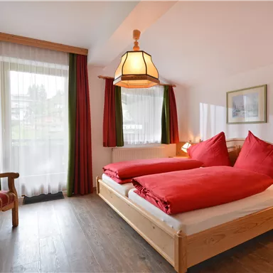 Ein gemütliches Zimmer mit zwei Betten und roten Bettdecken. Es gibt einen Sessel und große Fenster mit hellem Vorhang.