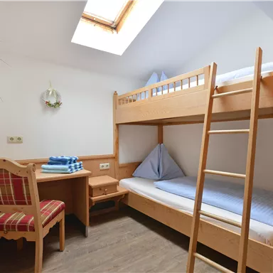 Ein einfaches Schlafzimmer mit einem Etagenbett und einem Tisch. Es gibt einen kleinen Stuhl und eine hohe Decke mit einem Fenster, das Tageslicht hereinlässt.