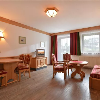 Ein einladendes Wohnzimmer mit Holzmöbeln und einer gemütlichen Sitzecke. Die Fenster lassen viel Licht herein und der Raum wirkt freundlich und warm.