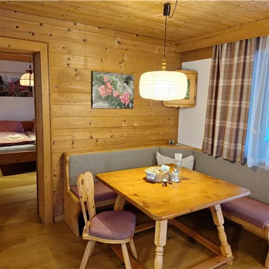 A cozy dining area with a wooden table and bench in a rustic room. In the background, there is a sleeping area and windows with curtains.