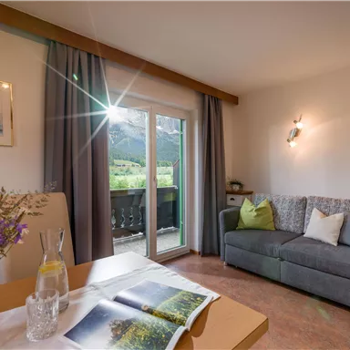 A cozy living room with a gray sofa and large windows overlooking a green landscape. On the table lies a book and a vase with fresh flowers.