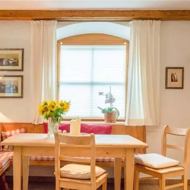 A cozy dining area with a wooden table and chairs. The walls are adorned, and there is a view of a sunny window seat with flowers.