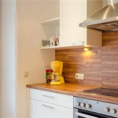 A modern kitchen with wooden shelves and a light color palette. The room is well-lit and features a stovetop and a coffee maker.