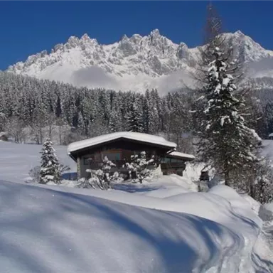 A cozy wooden house stands in a snowy landscape. In the background, impressive mountains rise under a clear blue sky.