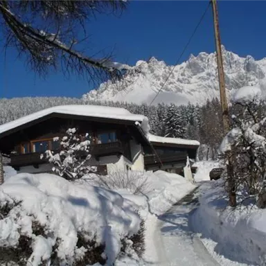 A charming house in the snow with surrounding trees. In the background, impressive mountains and a clear blue sky can be seen.