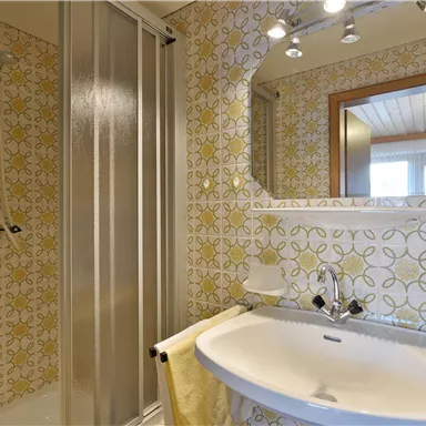 Ein Badezimmer mit einem Waschbecken, einem Spiegel und einer Dusche. Die Wände sind mit einem bunten, blumigen Muster gefliest.