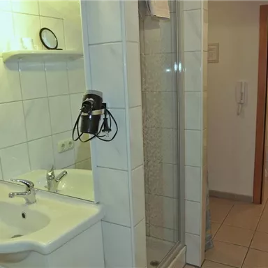 Ein einfaches Badezimmer mit einem Waschbecken, einem Spiegel und einer Dusche. Im Hintergrund ist eine getrennte Toilette zu sehen.