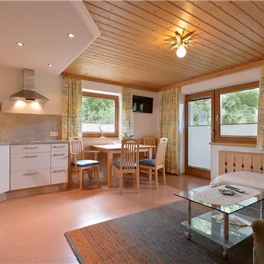 Eine moderne, gemütliche Wohnküche mit einer Sitzgruppe und einem Tisch. Große Fenster lassen viel Licht herein und bieten einen Blick nach draußen.