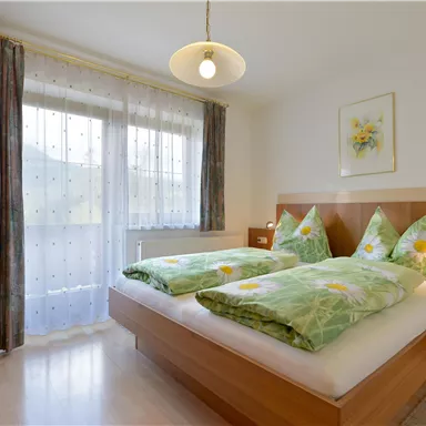 Ein helles Schlafzimmer mit zwei separaten Betten und bunten Bettdecken. Große Fenster sorgen für viel Tageslicht.