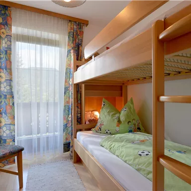 Ein gemütliches Kinderzimmer mit einem Etagenbett und bunten Vorhängen. Ein Stuhl steht neben dem Fenster, das viel Licht hereinlässt.