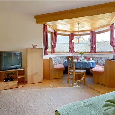 Ein gemütlicher Wohnraum mit einer Sitzecke und großen Fenstern. Der Raum ist mit Holzmöbeln und einem Fernseher ausgestattet.