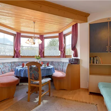 Eine gemütliche Essecke mit einem runden Tisch und gepolsterten Bänken. Große Fenster mit Vorhängen lassen viel Licht herein und schaffen eine einladende Atmosphäre.