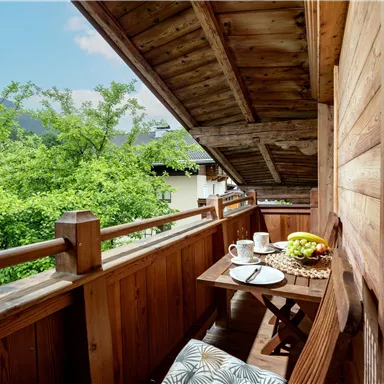 A cozy balcony with wooden paneling and a view of green trees. On the table are a cup of coffee and a fruit plate.