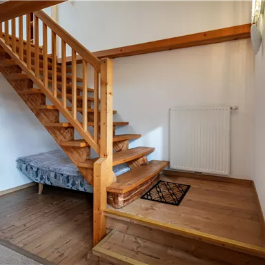 A bright hallway with a wooden staircase and a cozy wicker chair. A rug lies on the floor next to a heater.