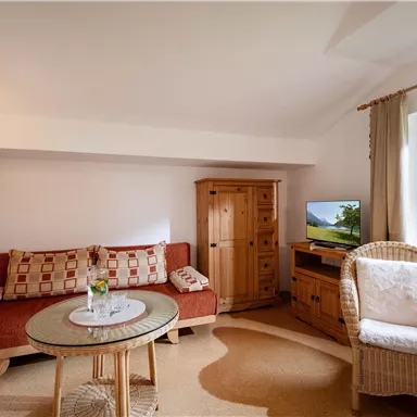 A cozy living room with a red sofa, a table, and a rocking chair. There is a window with a view outside and a television on a wooden cabinet.