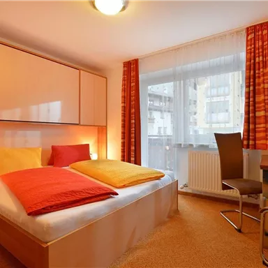 A cozy bedroom with a double bed and colorful pillows. A table and a chair are located next to the window, which lets in a lot of natural light.