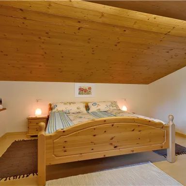 Ein gemütliches Schlafzimmer mit einem Holzbett und schrägen Decken. Neben dem Bett befinden sich Nachttische mit sanften Lampen.