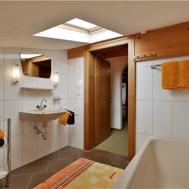 Ein modernes Badezimmer mit einer Badewanne und einem großen Fenster. Die Wände sind weiß gefliest, und orangefarbene Handtücher und Dekorationen setzen Akzente.