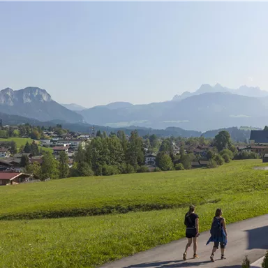 Eine idyllische Landschaft mit sanften Hügeln und einem Blick auf die Berge. Zwei Personen spazieren in der Nähe eines kleinen Dorfes.