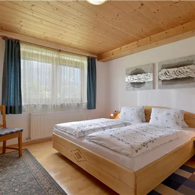 Ein gemütliches Schlafzimmer mit zwei Betten und Holzdecke. Vor einem Fenster stehen ein Stuhl und eine Lampe auf einem kleinen Tisch.