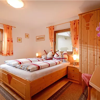 A cozy bedroom with a double bed and colorful pillows. The walls are light, and there are windows with curtains as well as a modern wooden dresser.
