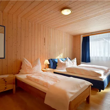 A cozy room with two single beds and wooden-paneled walls. The window lets in natural light and offers a view outside.