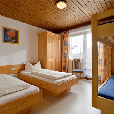 Ein einfaches Schlafzimmer mit zwei Einzelbetten und einem Etagenbett. Der Raum hat helle Holzverkleidungen und große Fenster mit Vorhängen.