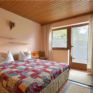 Ein gemütliches Schlafzimmer mit einem Doppelbett und bunten Bettdecken. Große Fenster geben einen Blick auf den Außenbereich und sorgen für viel Tageslicht.