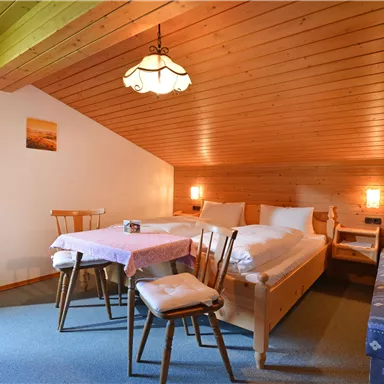 Ein gemütliches Zimmer mit Holzdecke, zwei Betten und einem Tisch mit Stühlen. Die Wände sind mit farbenfrohen Vorhängen und einem kleinen Bild dekoriert.