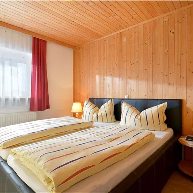 A cozy bedroom with wood paneling and a large double bed. Red curtains and a bedside table complement the peaceful atmosphere.