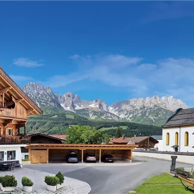Ein traditionelles Holzhaus neben einer Kirche, umgeben von grünen Wiesen und Bergen im Hintergrund. Der Himmel ist klar und blau.