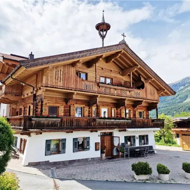 Ein traditionelles Holzhaus in den Alpen mit großer Veranda und Balkonen. Im Hintergrund sind majestätische Berge und eine klare Wetterlage zu sehen.