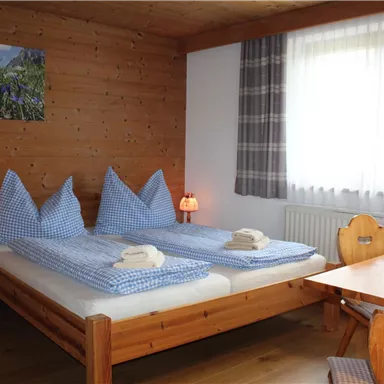 A cozy bedroom with two beds and checked bed covers. The room has wooden paneling and a table with flowers.