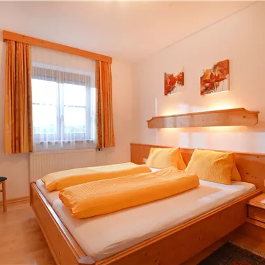 Ein gemütliches Schlafzimmer mit einem Doppelbett und zwei Kissen. Der Raum hat ein Fenster, Vorhänge in warmen Farben und einen Stuhl in der Ecke.