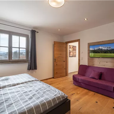 Ein gemütliches Schlafzimmer mit einem Doppelbett und einem lila Sofa. An der Wand befindet sich ein Fernseher und die Fenster bieten viel Tageslicht.