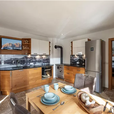 Eine moderne Küche mit Holzschränken und einem Esstisch. Im Hintergrund sind Geräte und eine gemütliche Atmosphäre zu sehen.