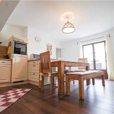 Eine helle Küche mit modernem Design und einem großen Esstisch aus Holz. Die Fenster lassen viel Tageslicht herein und sorgen für eine freundliche Atmosphäre.