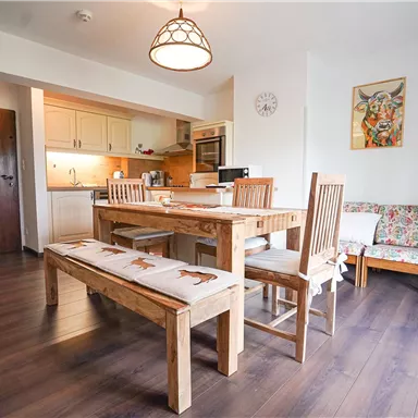 Eine moderne Küche mit einem Holztisch und Stühlen. Der Raum ist hell und freundlich, mit einem Teppich und Kunstwerken an den Wänden.