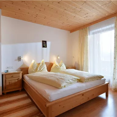 Ein gemütliches Schlafzimmer mit Holzdekor. Es gibt ein großes Bett, zwei Kissen und einen Stuhl neben einem Fenster mit Vorhängen.