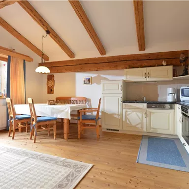 Eine moderne Küche mit Holzbalken und einem Esstisch. Das Zimmer ist hell und freundlich, mit einem Fenster, das auf die Berge blickt.