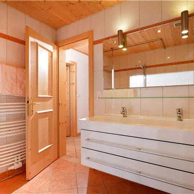 Ein modernes Badezimmer mit einem großen Spiegel und einem doppelten Waschbecken. Helle Fliesen und eine warme Holzdecke schaffen eine einladende Atmosphäre.