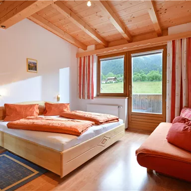 Ein gemütliches Schlafzimmer mit Holzdecke und einem großen Fenster. Das Zimmer verfügt über ein Doppelbett mit orangefarbenen Kissen und eine Sitzecke.