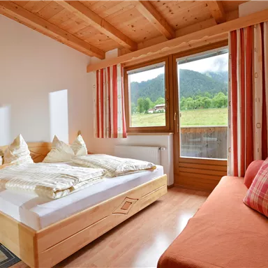Ein gemütliches Zimmer mit einem Doppelbett und einem Tagesbett. Große Fenster bieten einen Blick auf die Berge und viel Tageslicht.
